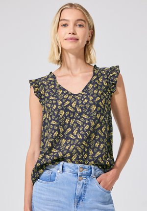 Kvinde med blondt hår iført en mørkeblå ærmeløs bluse med gult blomsterprint og lyseblå højtaljede jeans.