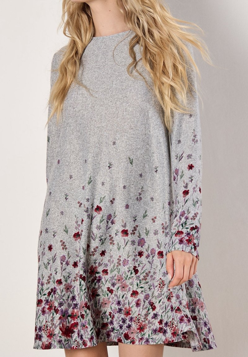 Femme portant une robe grise à manches longues avec un motif floral concentré près de l'ourlet et des manches.