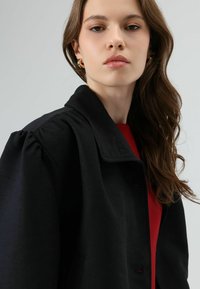 Zwarte jas met een gestructureerde kraag, pofmouwen en knoopsluiting aan de voorkant, gedragen over een rood topje. Soepele textuur en minimale ontwerpdetails.