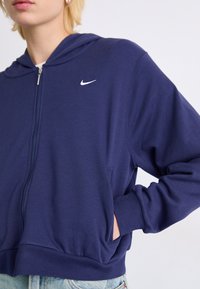 Sudadera con capucha azul marino con cremallera, confeccionada en tela suave, con bolsillo frontal, puños acanalados y pequeño logo blanco de Nike en el pecho.