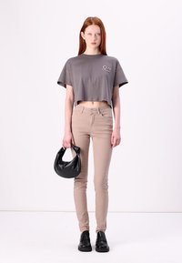T-shirt grise coupée avec logo, pantalon beige ajusté, sac à main noir et chaussures noires. Design simple avec un look décontracté et une texture lisse.