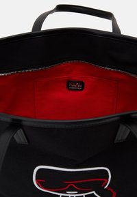 Svart tote-väska med läderhandtag, röd inredning och en logotyp-patch. Har en dragkedja och en broderad grafik p å framsidan.
