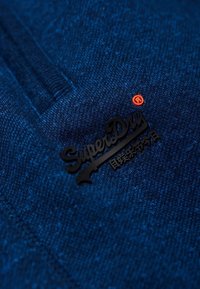 Gros plan sur un tissu bleu texturé avec un logo "SuperDry" noir et un petit symbole rouge de marque déposée sur le vêtement.