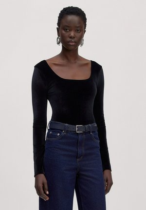 Frau trägt langärmliges schwarzes Samtoberteil mit eckigem Ausschnitt, dunkelblaue Jeans, schwarzen Gürtel und große silberne Creolen, steht vor schlichter Hintergrund.