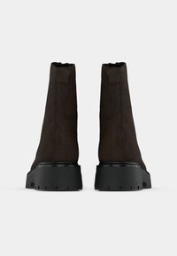 Brown suede ankle boots with a high shaft, black rubber sole, and textured accents at the heel. Features a smooth finish and sturdy design.