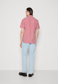 Chemise à manches courtes en vichy rouge et blanc avec un col, portée avec un jean bleu clair et des chaussures noires, avec un détail de pli au dos.