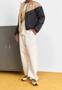 Chaqueta acolchada con parte superior en color marrón claro y sección inferior negra, camiseta crema, pantalones anchos crema y zapatillas beige con detalles oscuros.