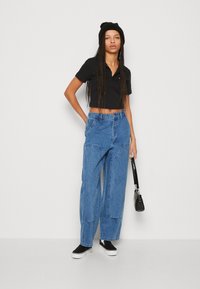 Črni skrajšani polo majica, svetlo modri široki traper hlače, črna kapa in črni športni copati. Model drži majhno črno torbico.