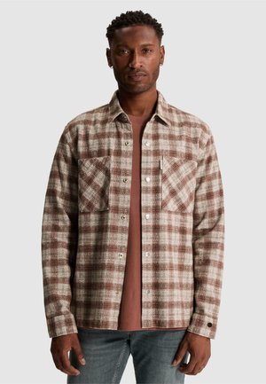 Homme aux cheveux courts et bouclés portant une chemise à carreaux beige et marron boutonnée sur un t-shirt couleur rouille et un jean bleu, debout devant un fond uni.