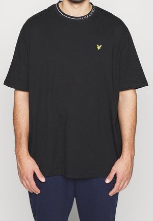 Uomo che indossa una t-shirt nera semplice a maniche corte con un piccolo logo giallo di un uccello sul petto sinistro e testo sul colletto.