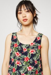 Top sem mangas floral com um fundo azul-marinho, flores vermelhas vibrantes e folhas verdes, feito de um tecido leve e com um decote redondo.