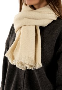 Sciarpa beige realizzata in tessuto testurizzato con un motivo a zig zag, con frange ai bordi, indossata sopra un indumento grigio scuro.