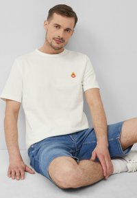 Weißes Baumwoll-T-Shirt mit rundem Halsausschnitt und kurzen Ärmeln, versehen mit einem kleinen gelb-roten Flammenmotiv mit der Aufschrift "BBQ". Blaue Jeansshorts.