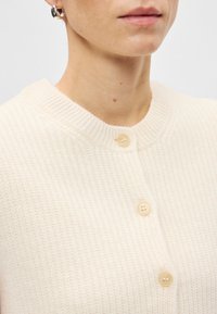 Ανοιχτό κρεμ πουλόβερ με υφή πλέξης, διαθέτει στρογγυλή λαιμόκοψη και τρία μπεζ κουμπιά μπροστά. Απλός και μίνιμαλ σχεδιασμός.