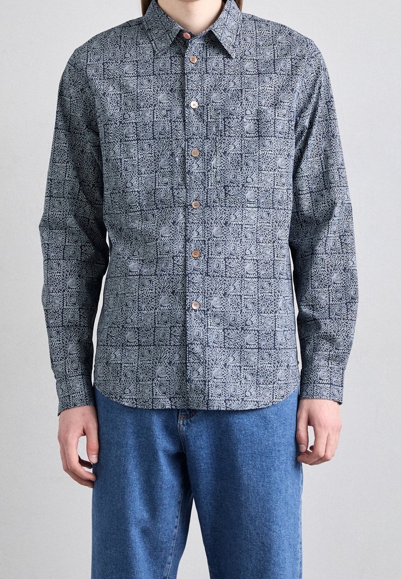 Langsleeve overhem met knopen in marineblauw met witte abstracte patronen, met een puntige kraag en bruine knopen. Gecombineerd met blauwe denim broek.