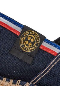 Tissu en toile bleu avec une étiquette tissée noire arborant un logo doré. Rayures rouges et blanches le long du bord. Couture en jute naturel visible.