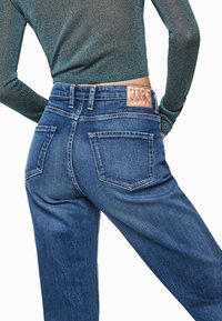 Vaqueros de mezclilla con un lavado azul oscuro, con cintura de talle medio, bolsillos traseros y una parche de cuero en la cinturilla. Textura suave.