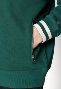 Sudadera verde con un bolsillo con cremallera en el lateral. Presenta puños acanalados con rayas blancas, textura suave y dobladillo ajustado.