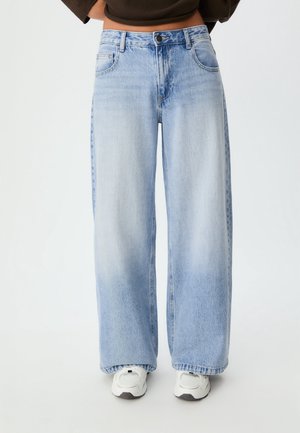 Personne portant un jean large bleu clair et des baskets blanches, debout devant un fond uni avec les mains sur les hanches.