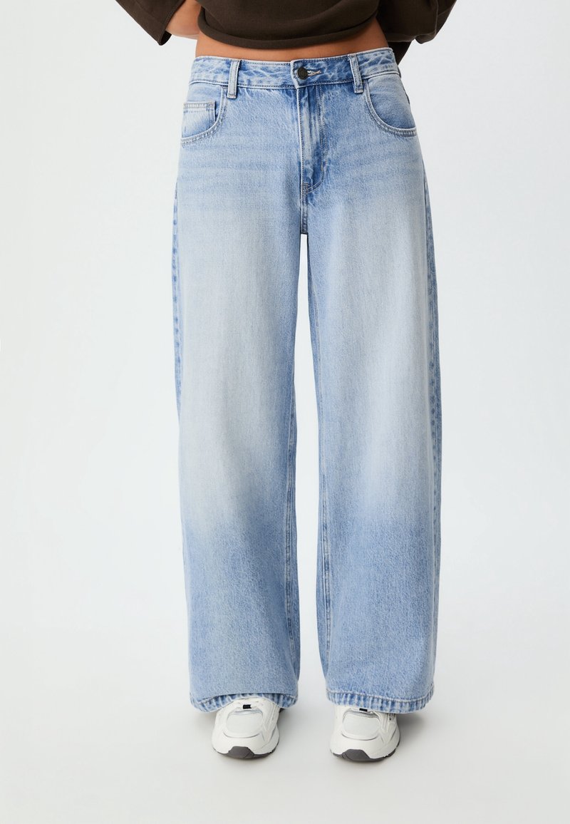Personne portant un jean large bleu clair et des baskets blanches, debout devant un fond uni avec les mains sur les hanches.
