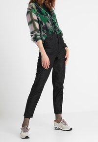 Kobieta w zielonej wzorzystej bluzce, czarnych dżinsach z wysokim stanem, pończoszkach w siatkę i białych trampkach, stojąca na jednolitym tle.