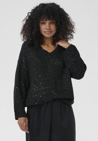 Vrouw met krullend haar, gekleed in een zwarte, met pailletten versierde V-halstrui en losse zwarte broek, staande tegen een effen lichte achtergrond.