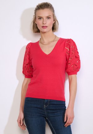 Žena v červenom tričku s výstrihom do V a čipkovanými nafúknutými rukávmi, modrých džínsoch, stojaca pred bielym pozadím tvárou v tvár.