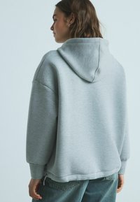 Grå hættetrøje med tekstureret stof, sænkede skuldre og sideslids, parret med højtaljede mørke denimjeans.