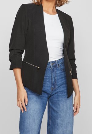 Frau trägt einen schwarzen Blazer mit Reißverschlusstaschen, weißes Oberteil und hoch taillierte blaue Jeans vor einem einfarbigen grauen Hintergrund.
