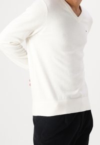 Muž v bílém svetr s výstřihem do V a dlouhými rukávy, v černých kalhotách, se rukou opřenou o dolní část zad, na jednobarevném pozadí.