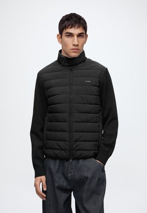 Ung mand iført en sort quiltet jakke med lynlås og høj krave samt mørke jeans, stående med den ene hånd i lommen mod en ensfarvet baggrund.