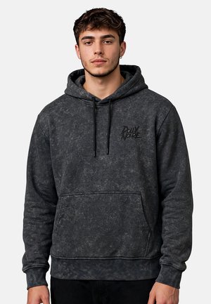 Joven con el pelo corto que lleva una sudadera stonewashed de color gris oscuro con bolsillo delantero y texto "Dolly Noire" en el pecho.
