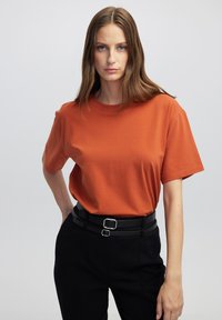 T-shirt a maniche corte e collo girocollo in tessuto arancione bruciato, abbinato a pantaloni neri a vita alta con cintura a doppia fibbia.
