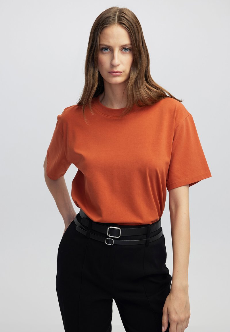 T-shirt a maniche corte e collo girocollo in tessuto arancione bruciato, abbinato a pantaloni neri a vita alta con cintura a doppia fibbia.