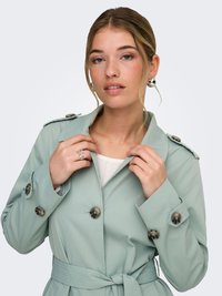 Trench-coat vert clair avec de gros boutons, une taille ceinturée et des épaulettes. Tissu lisse, manches longues et coupe ajustée.