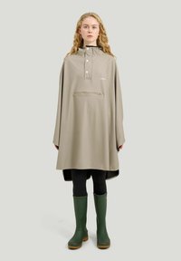 Kvinde iført en beige vandtæt poncho med frontlynlåslomme, sorte leggings og grønne gummistøvler, stående på en ensfarvet baggrund.