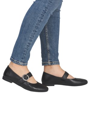 Personne portant des chaussures Mary Jane en cuir noir avec une boucle ronde argentée et un jean en denim bleu sur fond blanc.