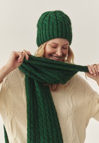 Berretto e sciarpa verdi lavorati a maglia con filato morbido e strutturato a motivo a coste. La sciarpa è avvolta intorno al collo sopra un maglione crema a trecce.
