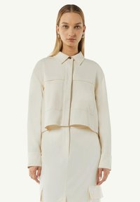 Przycięta, kremowa koszula z guzikami, z dwiema kieszeniami z przodu, długimi rękawami i klasycznym kołnierzykiem, wykonana z gładkiego materiału.