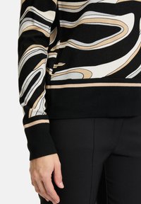 Schwarzer Pullover mit wellenförmigen Mustern in Beige, Weiß und Grau. Verfügt über lange Ärmel mit kontrastierenden Bündchenstreifen, kombiniert mit schwarzen Hosen.