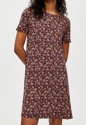 Femme portant une robe à manches courtes, au-dessus du genou, avec un motif floral foncé en rose, orange et vert sur un fond marron rougeâtre.