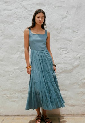 Frau mit langen dunklen Haaren, die ein ärmelloses, stufiges Maxikleid in Blau und Sandalen trägt, steht vor einer strukturierten weißen Wand.