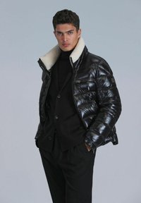 Black puffer jacket with a glossy finish, featuring a cream faux shearling collar and zippered front pockets. Worn over a black sweater.