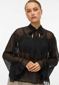 Černá průhledná blůza s límcem na mašličku, rozšířenými rukávy a volánkovými manžetami. Materiál má lehkou, průsvitnou texturu.
