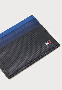 Két színű pénztárca, bordázott fekete textúrával, sima kék és tengerészkék kártyatartókkal, a sarokban egy kis logóval kiemelve.