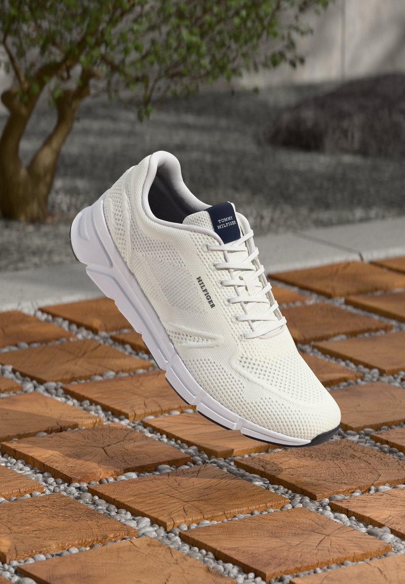 White knitted Tommy Hilfiger sneaker with lace-up design displayed above wooden tiles and small gray stones outdoors.