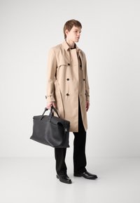 Trench coat bej cu un design dublu, nasturi negri; geantă de piele neagră tip duffel cu mânere rezistente; pantaloni și pantofi negri uni.