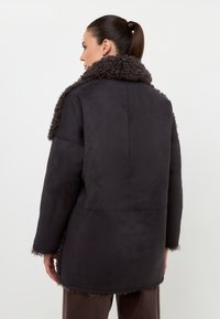 Abrigo de ante negro con un cuello de piel sintética texturizado y oscuro. Corte holgado, mangas largas y costuras visibles. Tela suave y textura blanda.
