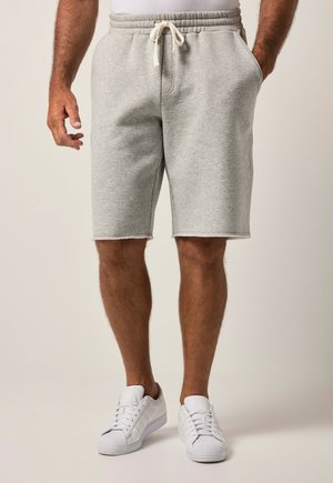 Mand iført lysegrå shorts med snøre og hvide sneakers, stående med den ene hånd i lommen mod en ensfarvet baggrund.