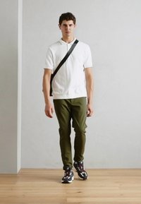 Hvid polo shirt, olivengrønne bukser og sorte og hvide sneakers. Modellen holder en sort skuldertaske, mens hun går på trægulv.